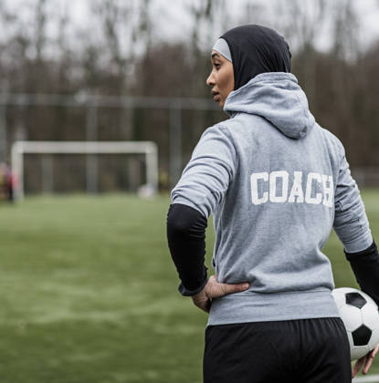 Jonge vrouwelijke voetbalcoach met hoofddoek op aan de rand van een voetbalveld